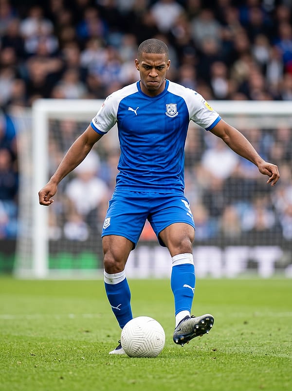 Player standing behind a football in a free kick stance, intense focus on the face, stadium goal and green pitch background.