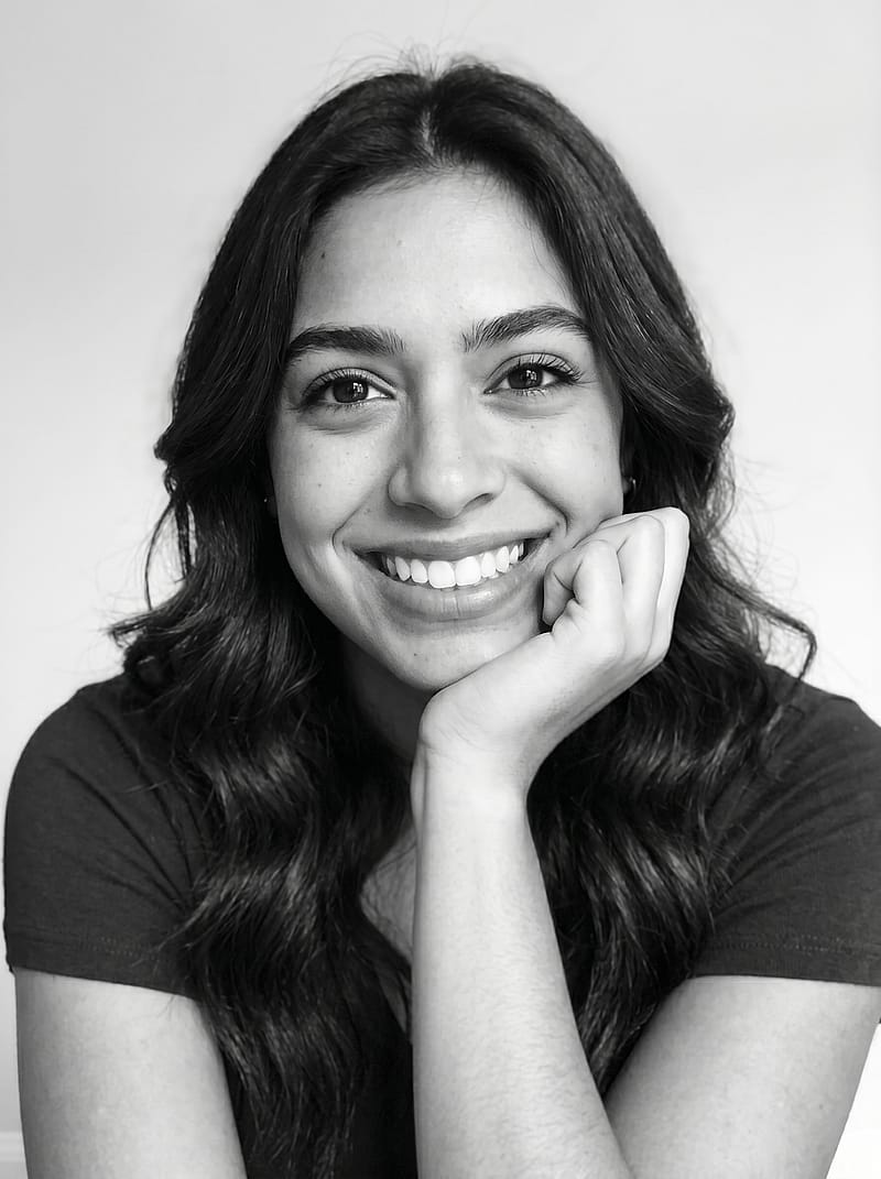 Joyful black and white studio portrait with a wide genuine smile, soft high-key lighting, and hand resting on chin.
