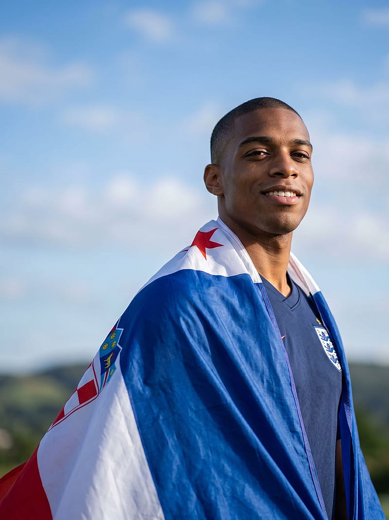 Fan wearing a national flag as a cape, standing proudly with wind blowing the fabric, blue sky and open air background.